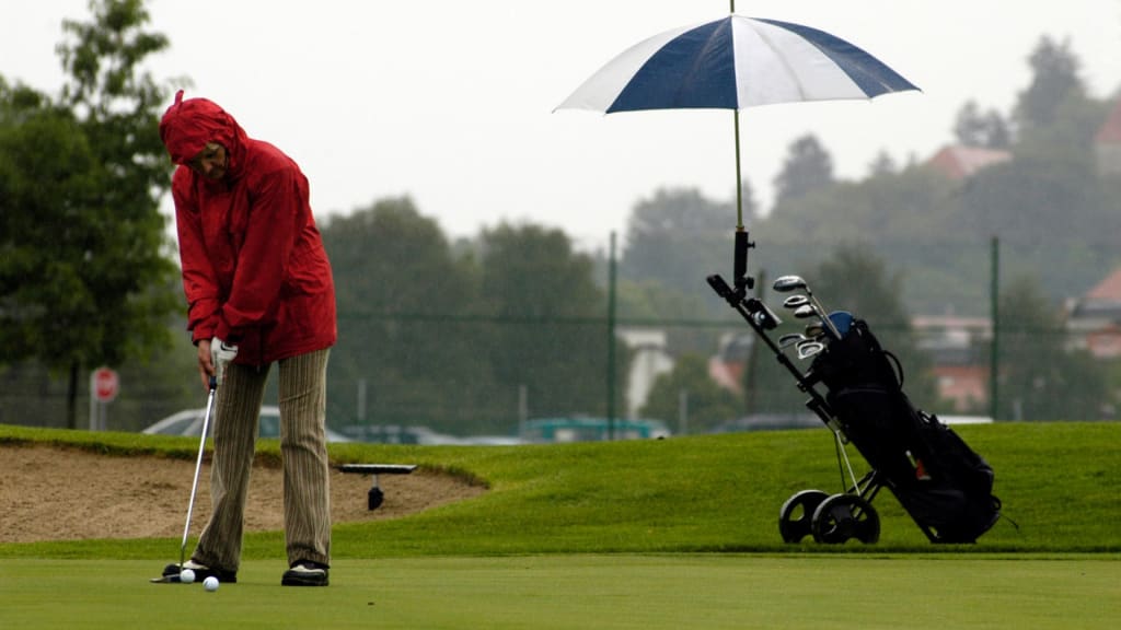 A person wearing a red rain jacket is putting on a golf green under cloudy skies. An umbrella is attached to their golf cart in the background.